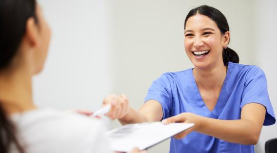 Young woman in scrubs hands other woman pen and notepad.